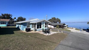 A small, single-story building with a covered outdoor seating area, a grassy yard, and a view of a calm body of water. - View of Lake Weir & private beach access  (Weirsdale)