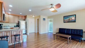 A spacious living area with a wooden floor, a black sofa, a wooden coffee table, and a ceiling fan. - Palm Desert Condo. Near Tennis Gardens, Public Golf, Dog Park. EZ on the wallet! (Palm Desert)