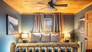 Une chambre à coucher au style rustique, avec un lit en bois, un ventilateur de plafond et une fenêtre agrémentée de rideaux.