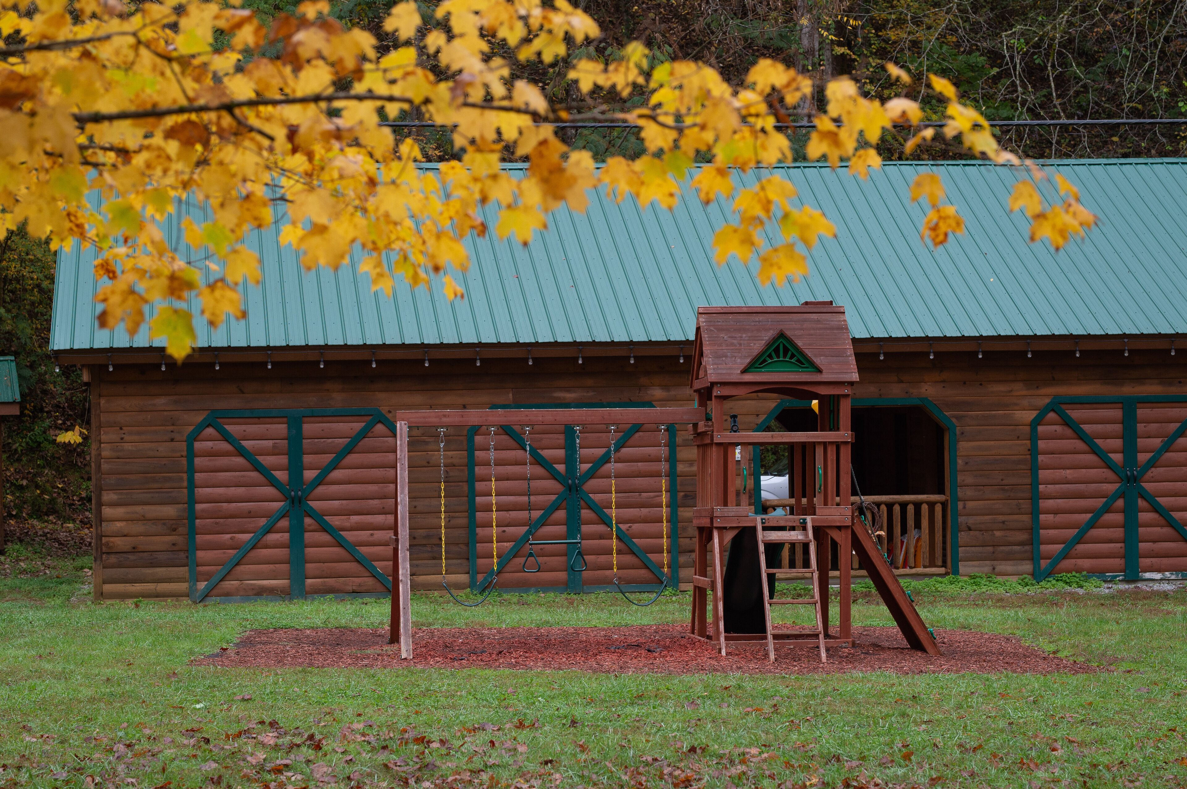 A wooden play structure with a swing and slide in front of a rustic building with a green roof and cross-braced walls.