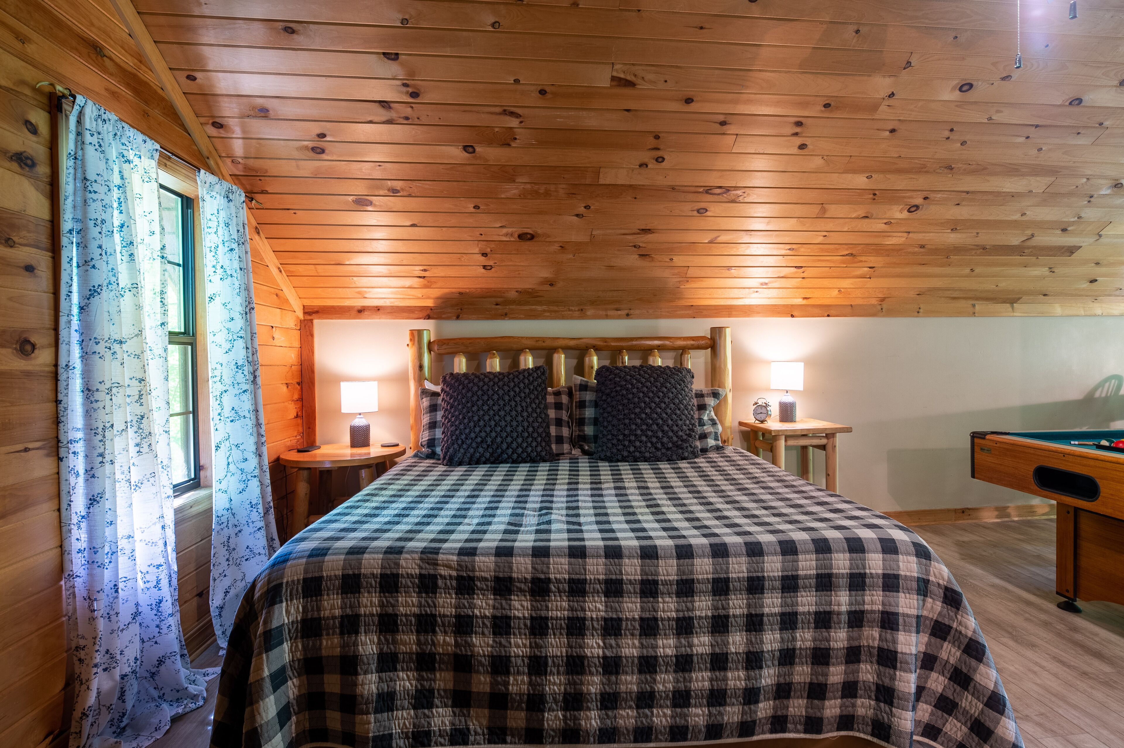 A cozy bedroom with a wooden ceiling, a checkered bedspread, two bedside lamps, and a pool table.