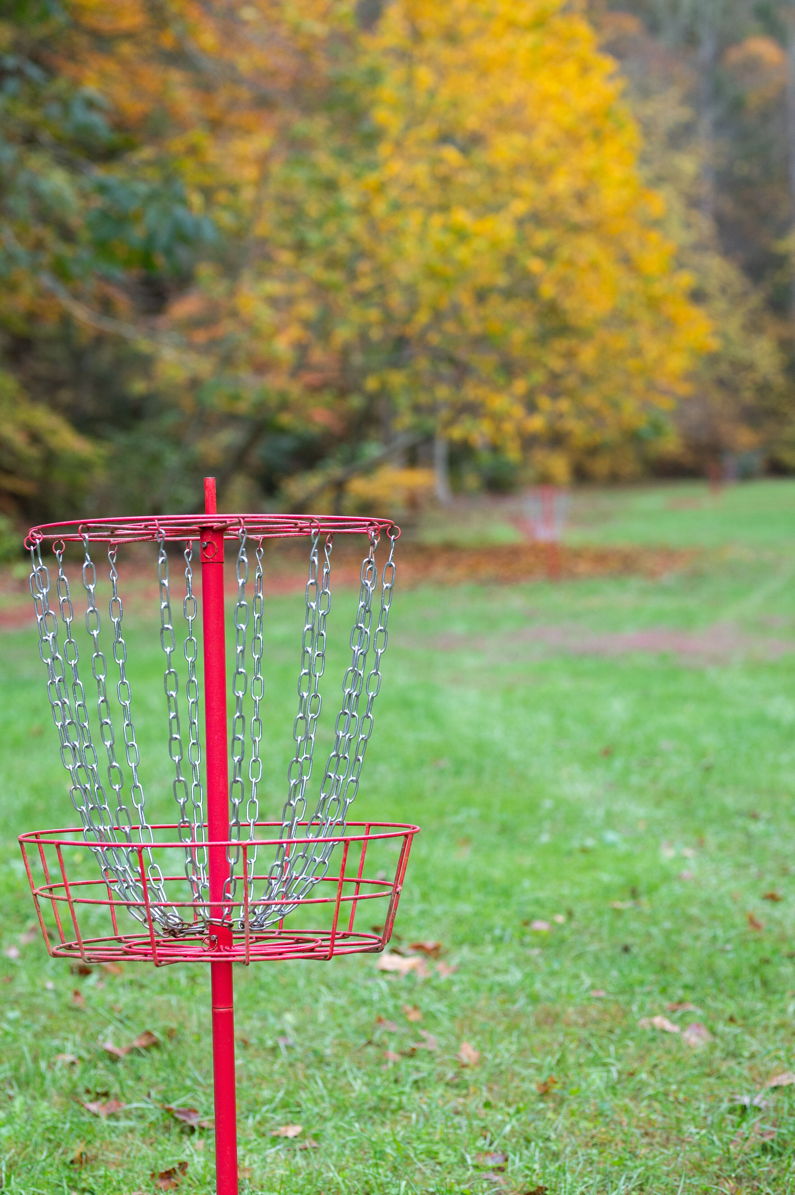 A red disc golf basket with metal chains, set against a grassy field with autumn-colored trees in the background.