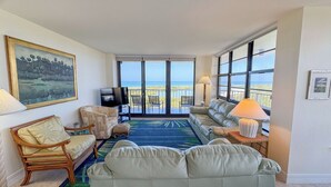 Ein geräumiger Wohnbereich mit großem Fenster zum Meer, einem bequemen Sofa, einem Fernseher und einem gerahmten Gemälde.