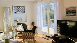 Ein Wohnzimmer mit Glastisch, Korbsessel, Sofa, Fernseher und einem großen Fenster mit Vorhängen.