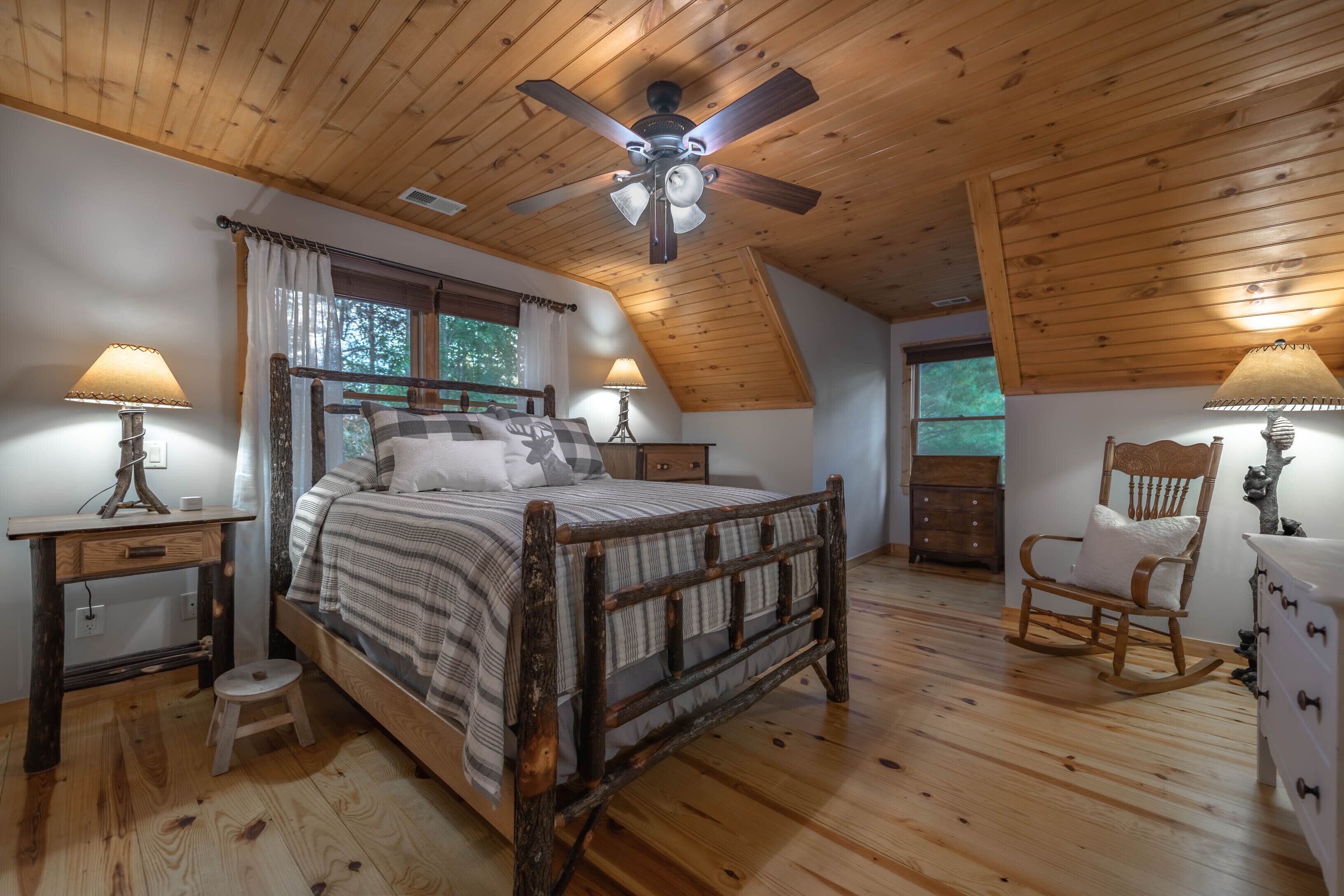 Une chambre à coucher avec un plafond en bois, un lit avec une tête de lit, deux tables de chevet avec des lampes, un fauteuil à bascule et une fenêtre avec des rideaux.