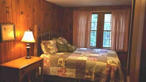 Ein Schlafzimmer mit Holzboden, einem Bett mit Steppdecke, einem Nachttisch mit Lampe und einem Fenster mit Vorhängen.