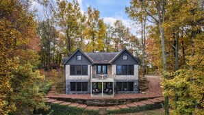 A two-story house with a balcony, surrounded by trees and a pathway leading to the entrance. - Modern Home with Mountain Views, Hot tub, Pool Table, and Gourmet Kitchen. (Tryon)