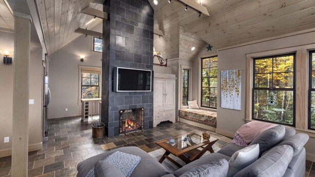 A spacious living room with a stone fireplace, a large gray sofa, and a view of a forest through large windows. - Modern Home with Mountain Views, Hot tub, Pool Table, and Gourmet Kitchen. (Tryon)