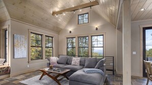 A spacious living room with a gray sectional sofa, a wooden coffee table, and large windows offering a view of the outdoors. - Modern Home with Mountain Views, Hot tub, Pool Table, and Gourmet Kitchen. (Tryon)