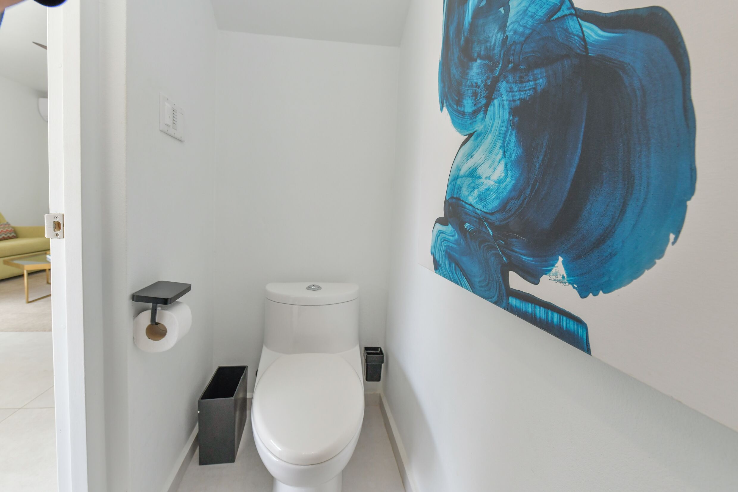 A bathroom with a white toilet, a trash bin, and a wall-mounted shelf.