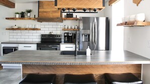 A modern kitchen with a large island, stainless steel appliances, wooden shelves, and a hanging light fixture.
