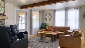 A living room with a brown leather sofa, a black leather chair, a wooden coffee table, and a patterned rug. There is a large wall clock and a plant in the corner.