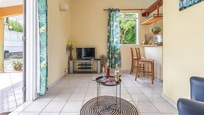 A living room with a TV, a dining area with chairs, a small table with drinks, and a view of greenery outside. - Cottage La Perle  ( pool, BBQ, view on moutains, sea, rivers, forests) (Basse-Terre)