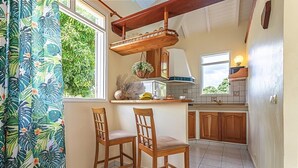 A kitchen with wooden cabinets, a hanging shelf, and two wooden chairs. - Cottage La Perle  ( pool, BBQ, view on moutains, sea, rivers, forests) (Basse-Terre)