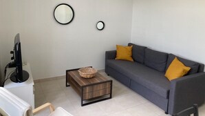 Un moderno salón con un sofá gris, dos almohadas color mostaza, una mesa de centro de madera con una cesta tejida, un televisor de pantalla plana sobre un soporte blanco y dos espejos colgados en la pared.