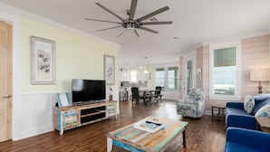 A spacious living room with hardwood floors, a ceiling fan, a television, a coffee table, a blue sofa, and a patterned armchair.