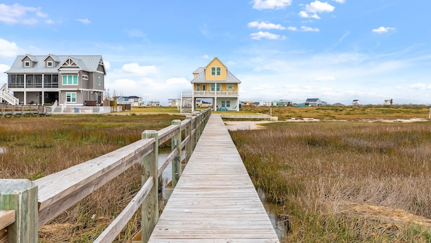 A wooden boardwalk leading to a yellow house with a balcony, surrounded by marshland and other houses.