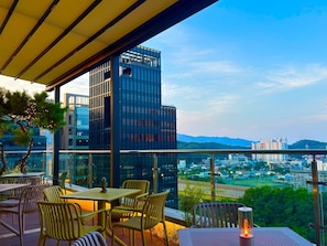 Un espacio al aire libre con mesas y sillas, con vistas a un paisaje urbano de edificios modernos y colinas distantes.