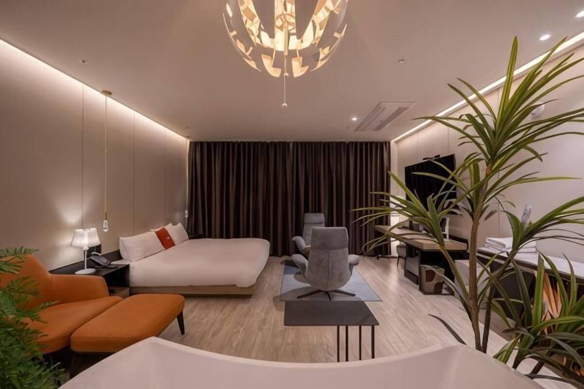A modern hotel room with a large bed, a desk, a chair, a TV, and a plant.