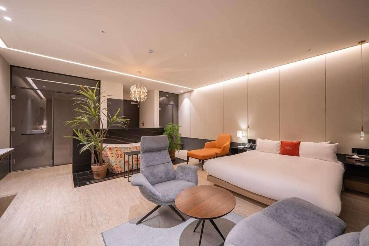 A modern hotel room with a large bed, two armchairs, a small round table, a desk, and a potted plant.