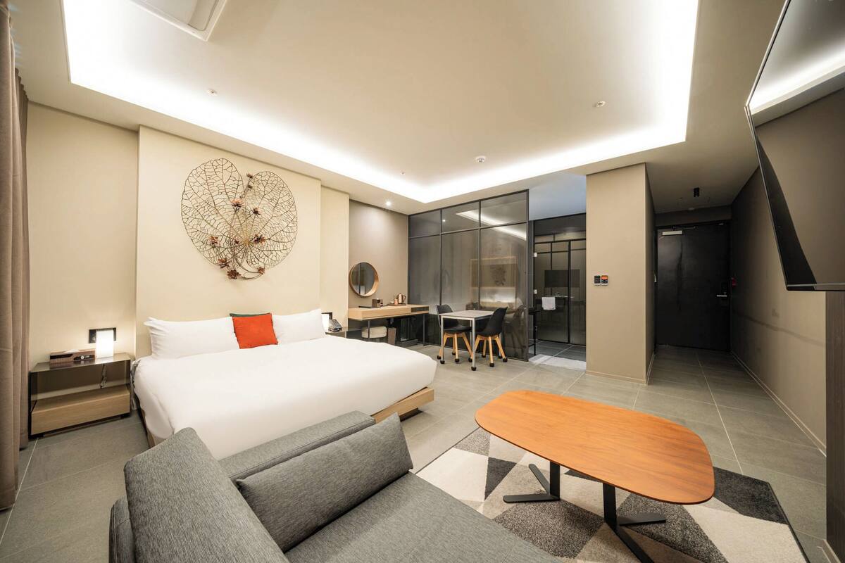 A modern hotel room with a large bed, a desk, a sofa, and a dining table.