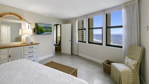Une chambre à coucher comprenant un lit, une chaise, un bureau avec une lampe, une fenêtre donnant sur l’océan et une porte menant à une autre pièce.