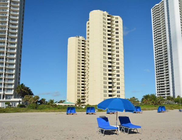 Ein Strand mit blauen Liegestühlen und einem blauen Sonnenschirm, umgeben von hohen Gebäuden.