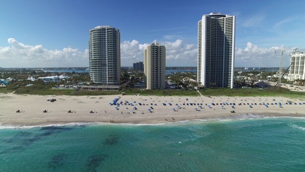 Un paysage urbain côtier avec trois immeubles de grande hauteur, une plage de sable bordée de nombreux parasols et une eau turquoise et limpide.