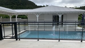 A swimming pool with a glass barrier, a covered pavilion, and a hilly landscape in the background. - Villa Saphir Equipée (Gourbeyre)