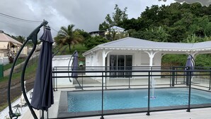 A swimming pool with a glass barrier, surrounded by a white gazebo and a white building. - Villa Saphir Equipée (Gourbeyre)