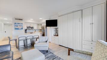 Un salon moderne avec un canapé, un fauteuil et un coin repas agrémenté de tabourets de bar. Au fond, on aperçoit une cuisine avec des meubles blancs et un téléviseur à écran plat fixé au-dessus du comptoir.
