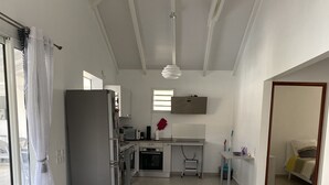 A modern kitchen with a refrigerator, oven, and sink. There is a dining area with a table and chairs, and a living room with a sofa. - Villa Rubis equipped for people with reduced mobility  (Gourbeyre)