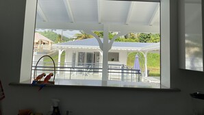 A room with a large window overlooking a covered outdoor area with a white structure and greenery. - Villa Rubis equipped for people with reduced mobility  (Gourbeyre)