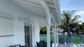 A covered outdoor area with a table and chairs, a covered walkway, and a view of a palm tree and house in the background. - Villa Rubis equipped for people with reduced mobility  (Gourbeyre)
