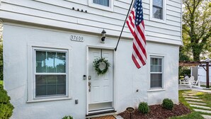 Een wit huis met twee verdiepingen, een voordeur, een nummerbord, een kleine tuin en een Amerikaanse vlag.