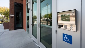 Entrada de un edificio con puerta de vidrio, señal de accesibilidad para sillas de ruedas y panel de control digital de ascensor.