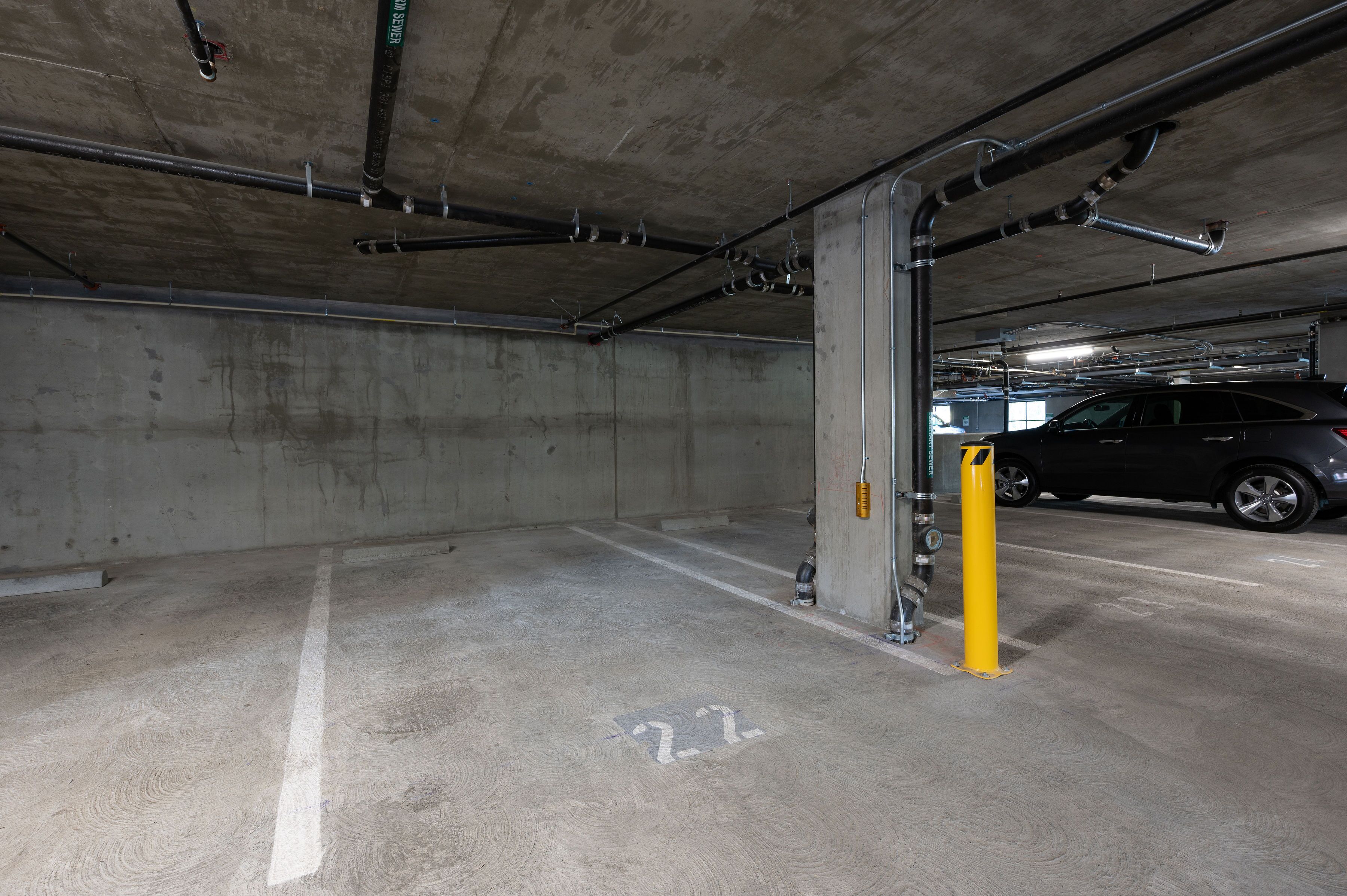 A parking garage with concrete walls and floors, marked parking spaces, and visible pipes and ducts.