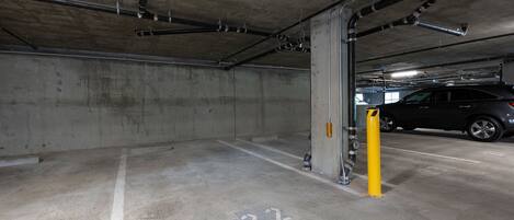 A parking garage with concrete walls and floors, marked parking spaces, and visible pipes and ducts.