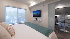 Una habitación de hotel moderna con un ventanal grande, una cama con manta verde, un televisor montado en la pared y una zona de kitchenette.