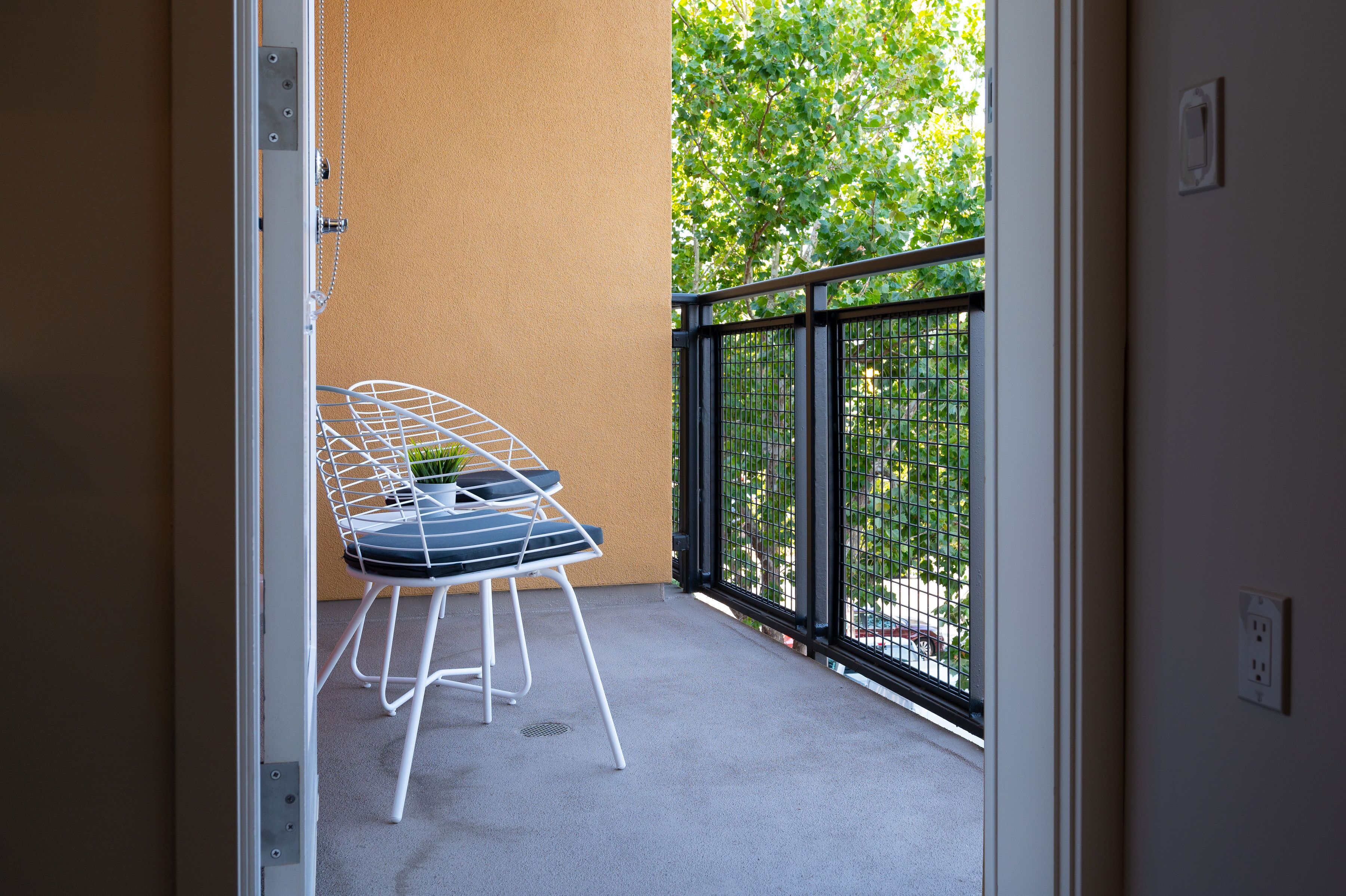 A balcony with a white metal chair and a small table, a beige wall, and a view of greenery through a black metal fence.