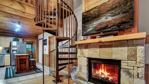 A cozy living room with a stone fireplace, a wooden spiral staircase, and a mounted TV. - Heated Rst Pool-Dollywood-WIFI -Arcade-Parkway-Families-Shopping-Hot Tub-Shows (Pigeon Forge)