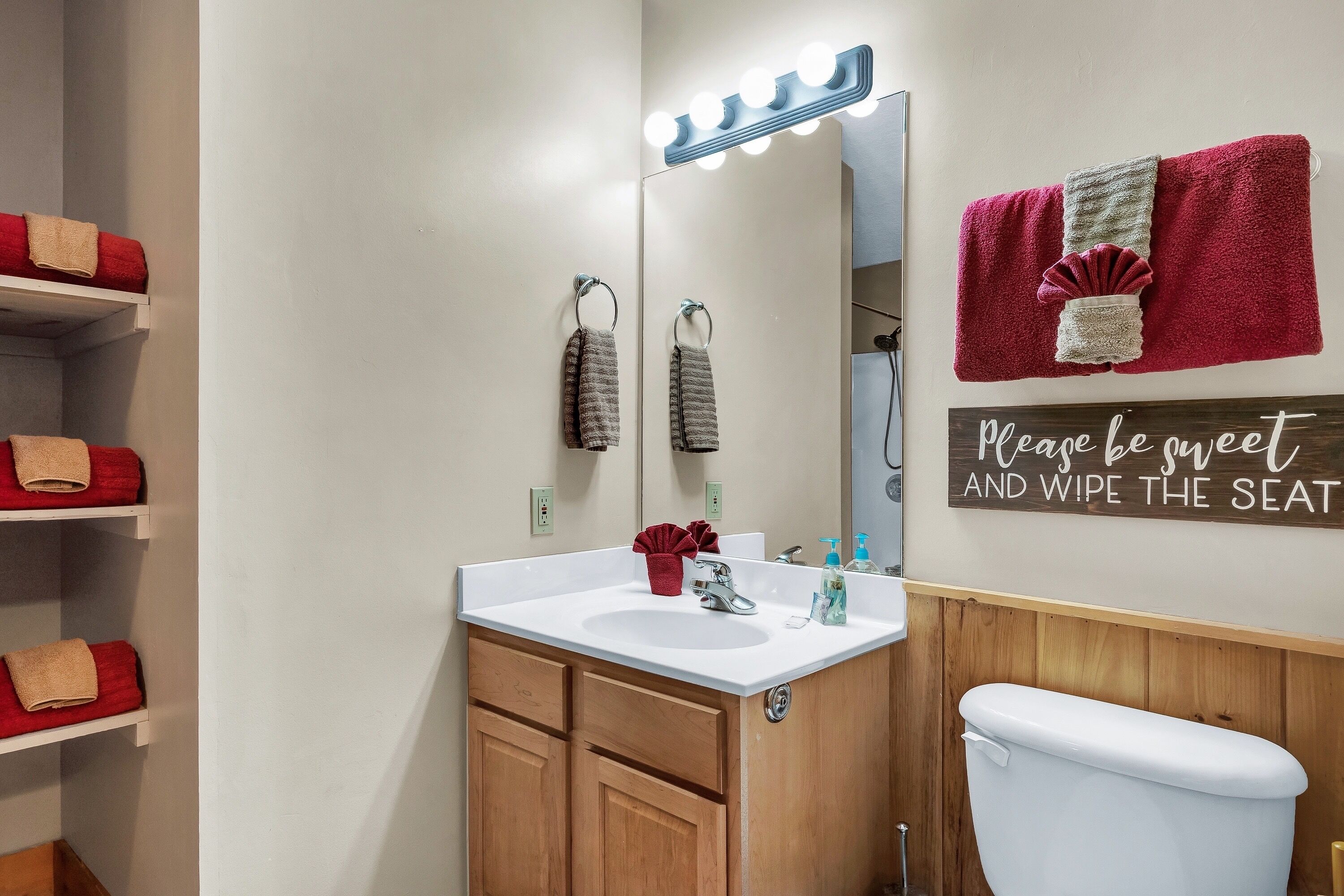 A bathroom with a vanity, sink, mirror, and toilet. There are towels on shelves and a sign on the wall.