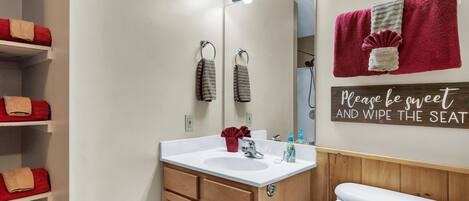 A bathroom with a vanity, sink, mirror, and toilet. There are towels on shelves and a sign on the wall.