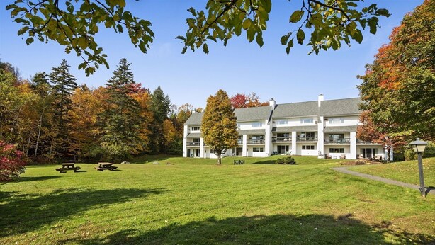 A multi-story building with a flat roof, surrounded by a well-maintained lawn and trees with autumn foliage. - Condo at Stowe`s Toll House | Ski-in/Ski-out! (Stowe)