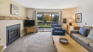 A living room with a fireplace, a television, a sofa, a coffee table, and a balcony with a view of trees and a fence. - Condo at Stowe`s Toll House | Ski-in/Ski-out! (Stowe)
