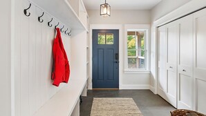 Un couloir avec une porte bleu foncé, des murs blancs, un porte-manteau suspendu avec une veste rouge, et un tapis tissé sur le sol.