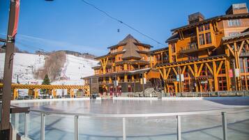 Un immeuble à plusieurs étages, avec un télésiège visible en arrière-plan, une patinoire couverte en verre au premier plan et une pente de montagne.