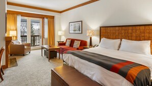 A hotel room with a large bed, a sofa, a desk, and a chair. There is a window with curtains, a framed picture on the wall, and a wooden headboard. - 2nd Floor Studio in The Lodge at Spruce Peak (Stowe)