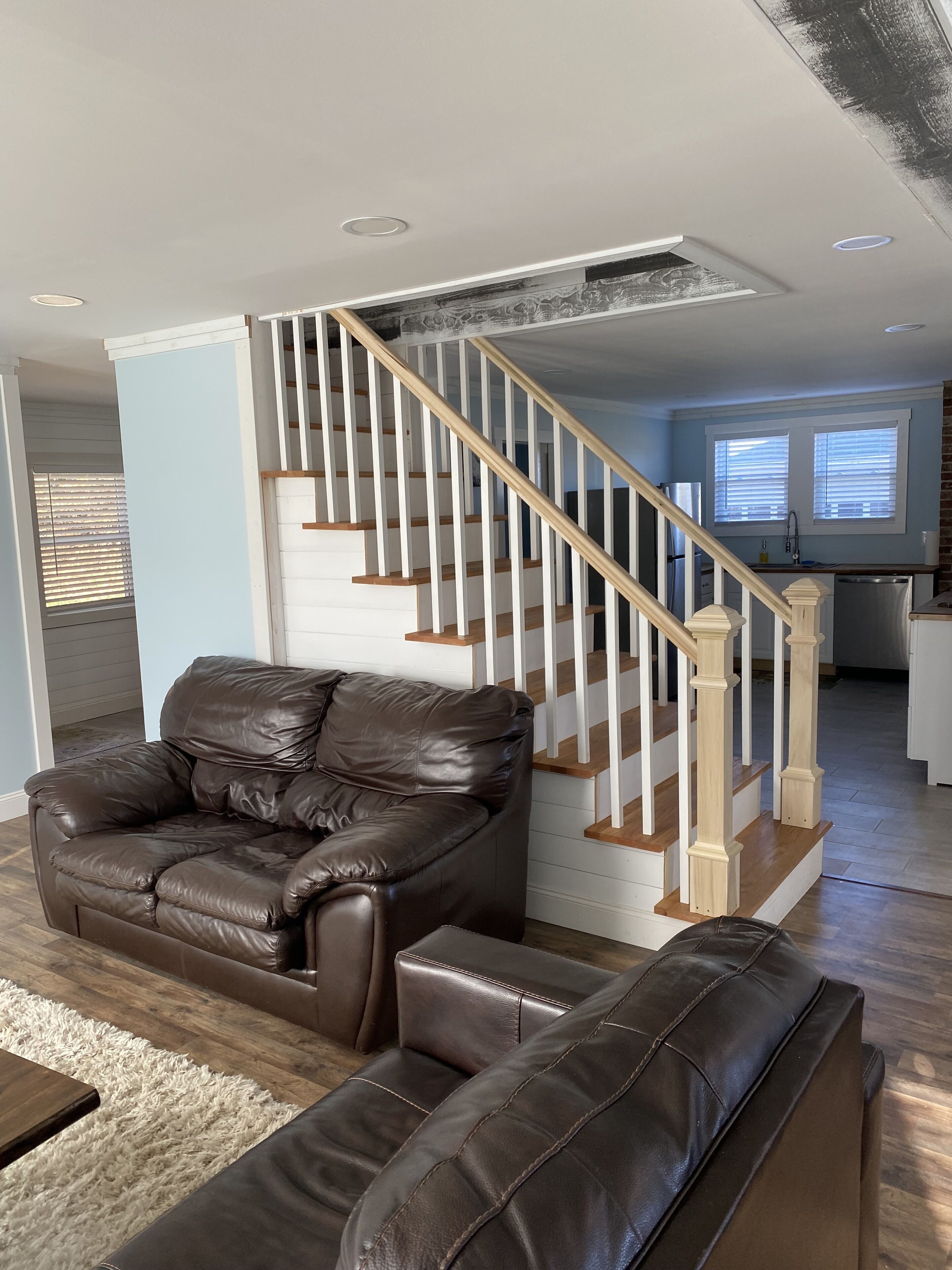 A modern living room with a brown leather sofa, a matching bench, and a wooden staircase with white balusters.