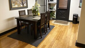 A dining area with a wooden table, chairs, a vase with flowers, a refrigerator, a microwave, and a waste bin.